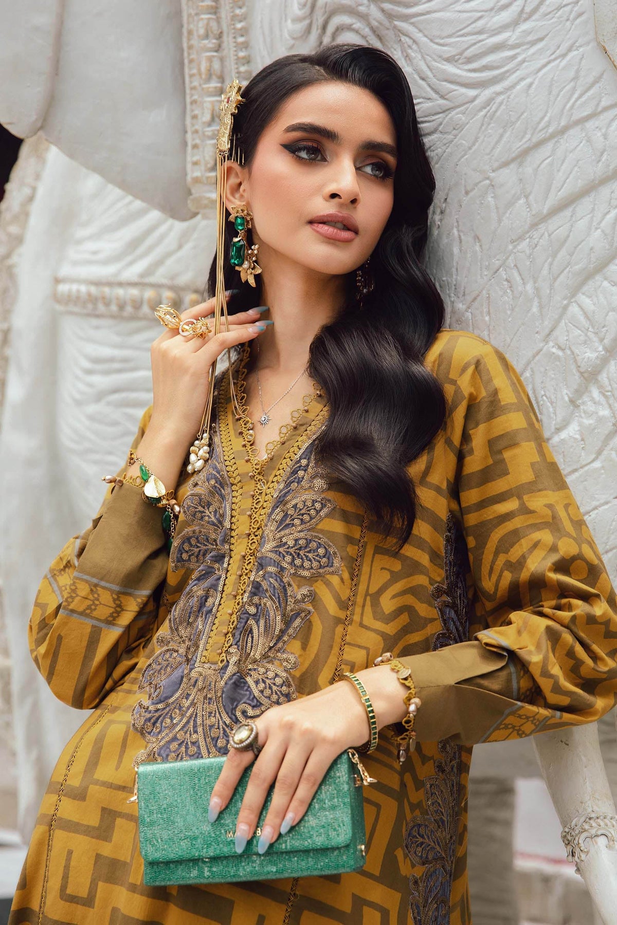 Woman in a mustard yellow and brown patterned outfit holding a green clutch against an elephant and architectural background.