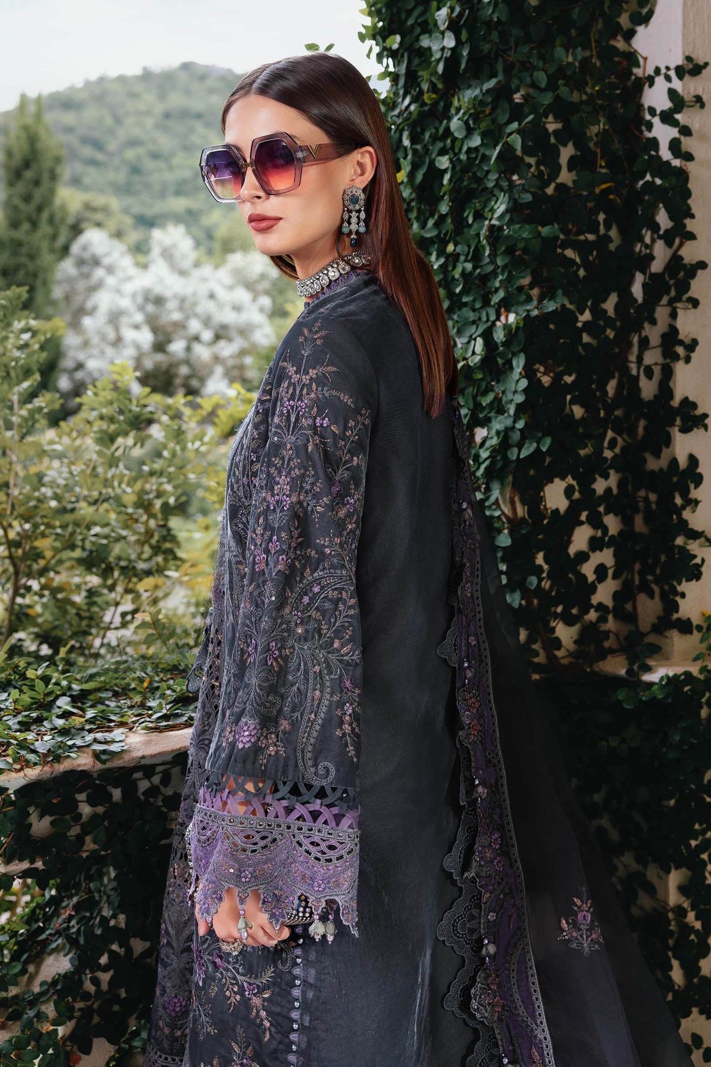 Woman in a dark outfit with floral patterns standing against a green leafy background