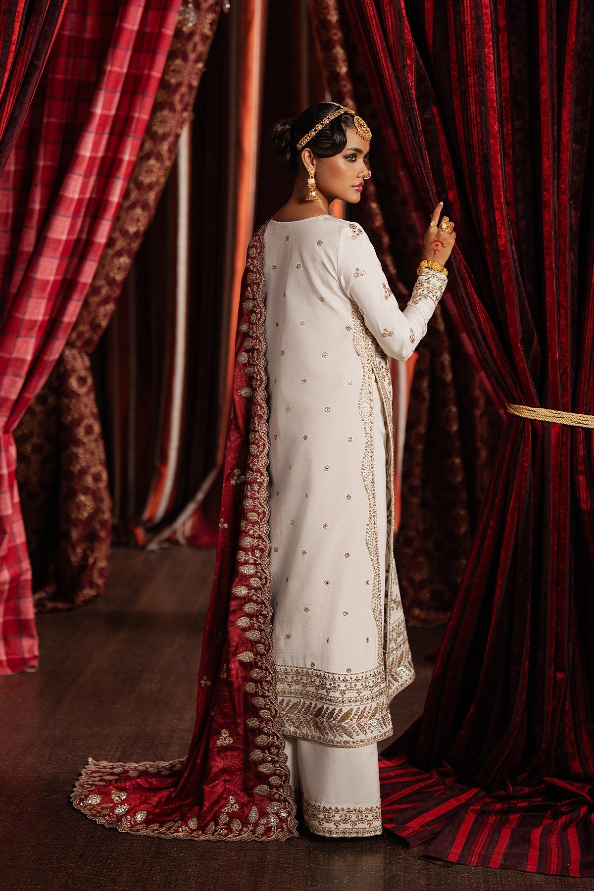 Woman in traditional white and gold outfit standing behind red curtains